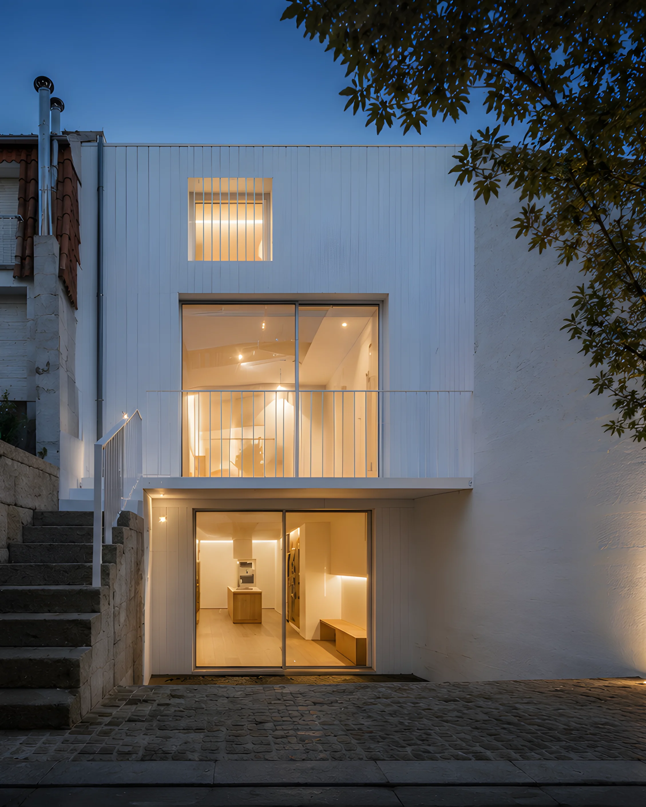 Fachada de vivienda estrecha rehabilitada, con nueva piel blanca, huecos verticales y acceso desde calle/patio empedrado.